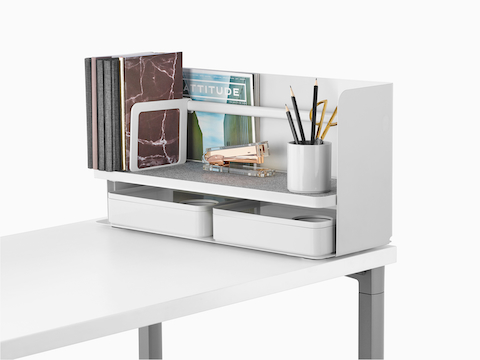 A small white Ubi desktop organiser with a grey non-skid shelf holds books, a stapler, a pencil cup, and two storage boxes.