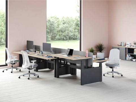 A bank of six Ratio desks with one positioned at a standing height. Three grey and white Lino chairs are set around the desks with a grey Paragraph storage unit in the background.