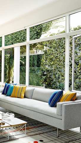 A cluster of Eames Wire Base Low Tables serves as a coffee table for a light gray Bolster sofa situated before a wall of windows.