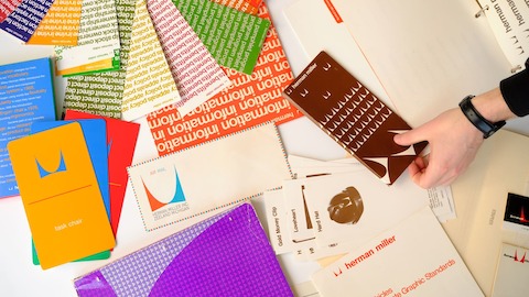 Colourful brochures from Herman Miller's archive are displayed loosely on a white tabletop with a hand picking up one.