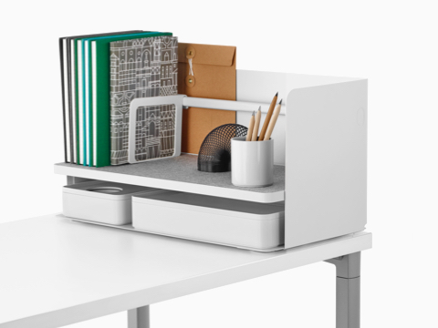 A large white Ubi desktop organiser with a grey non-skid shelf holds books, a pencil cup, and two storage boxes.