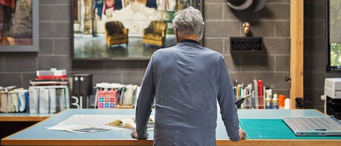 Designer Don Chadwick, his back to the camera, stands at a tall drafting table with a few books and cutting boards and bookshelf behind, in his studio.