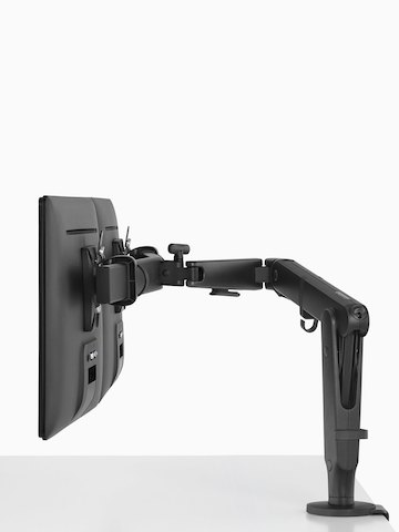 A side view of two monitors suspended above a white work surface from a black Ollin Monitor Arm.