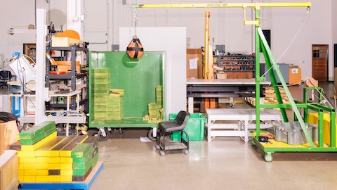 A brightly colored testing facility with green and yellow carts and piles of wood, with a basketball hanging from a yellow crane to act as a weighted wrecking ball.